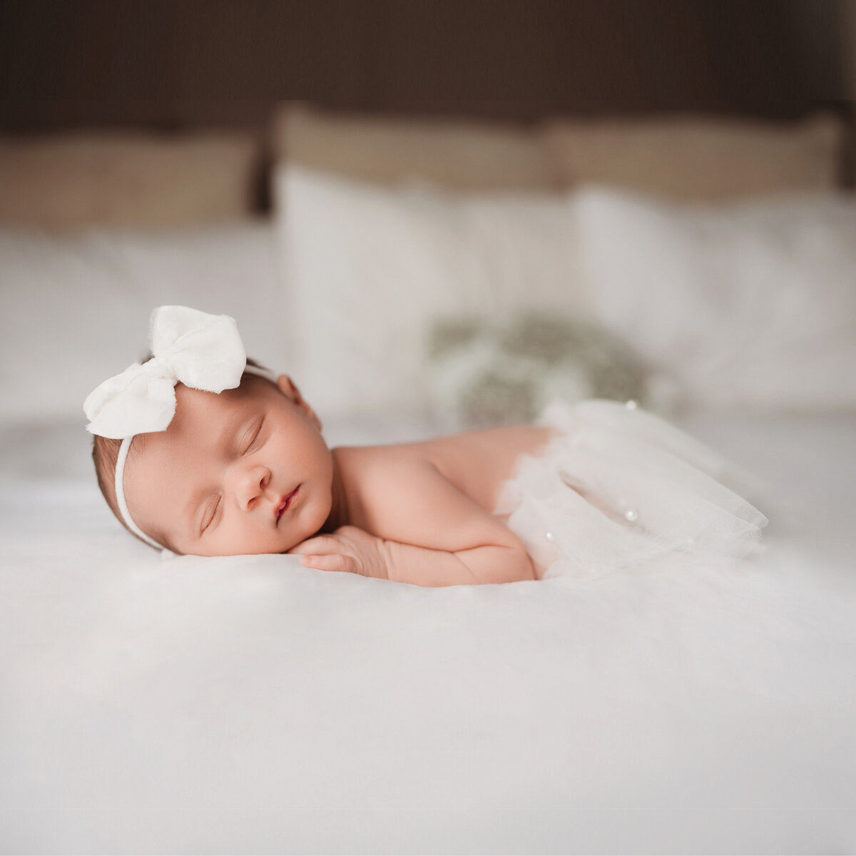 newborn baby girl in white bow and white pearl tutu laying on white bed on her tummy