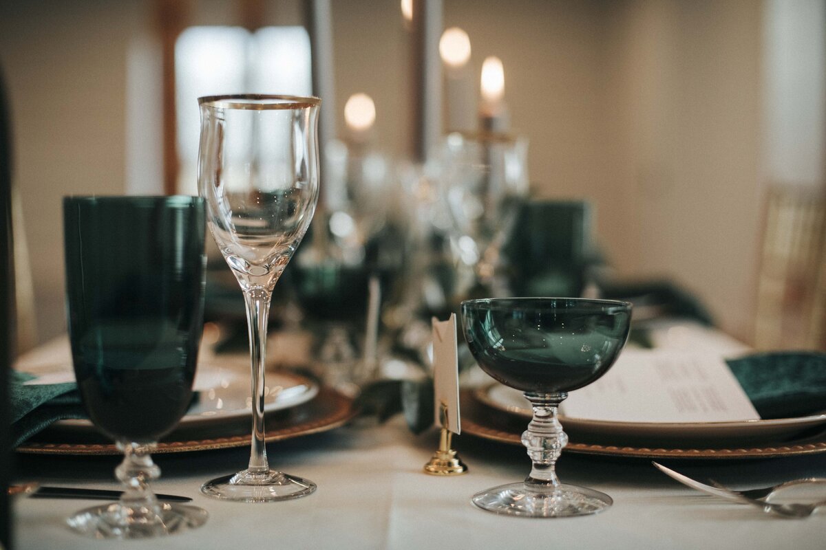 White squared place cards set on a reception table with dark green glasses, napkins, white plates and gold chargers.