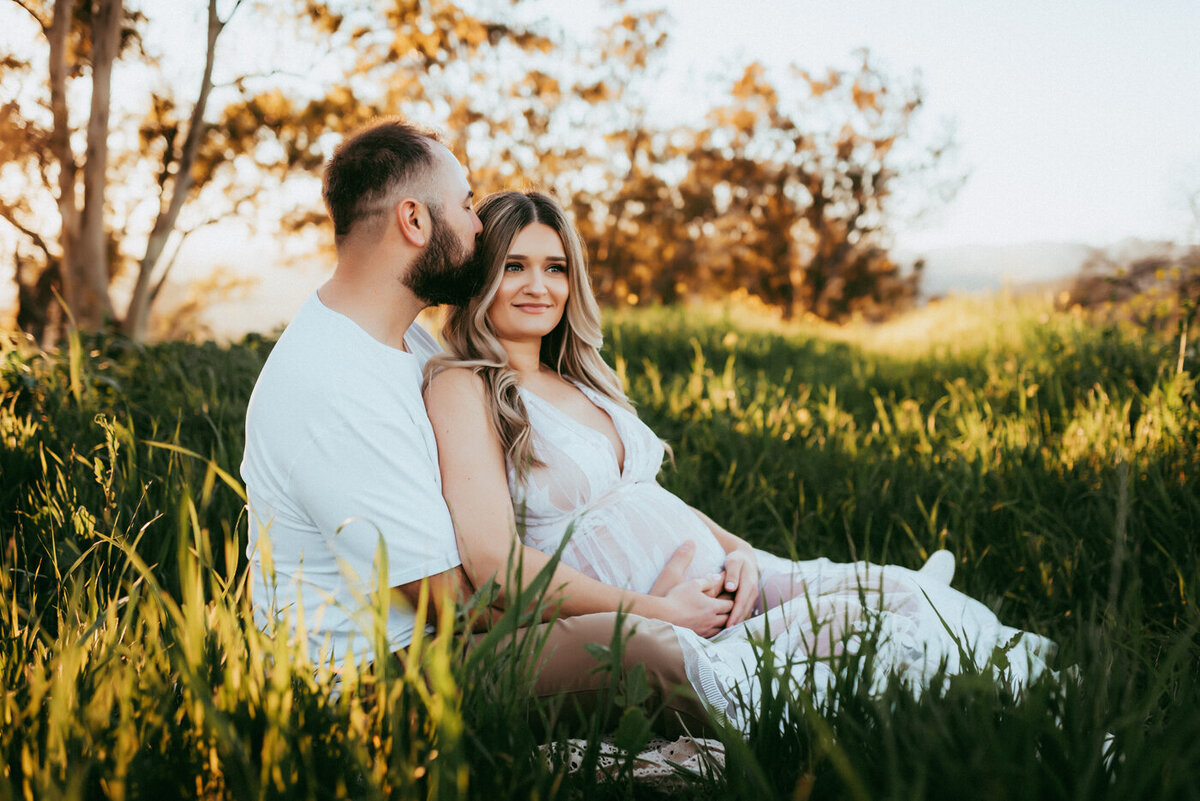 Pregnant mama and husband sitting down in a field of green grass