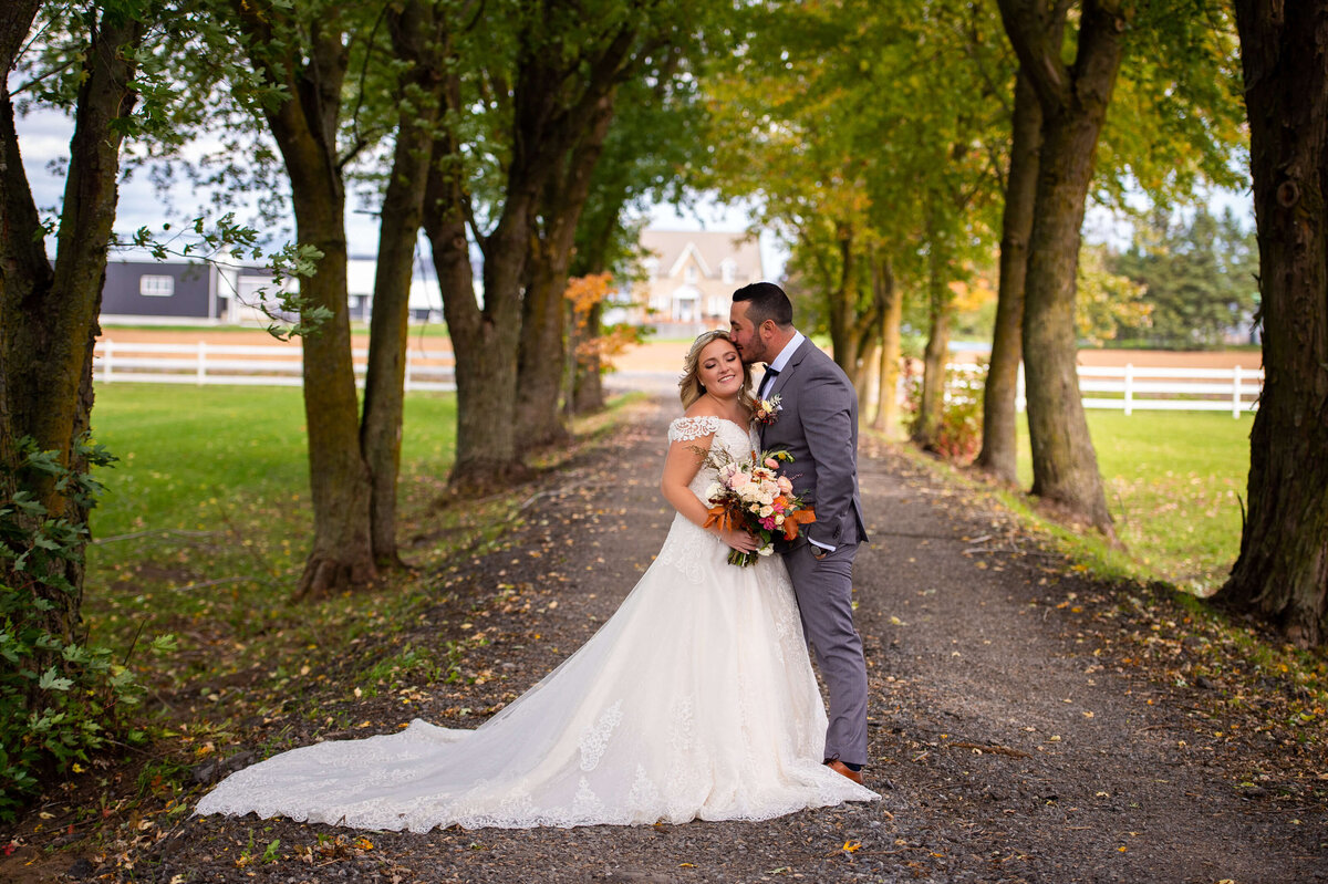 a groom in a grey suit kissing his bride on the cheek surrounded by fall coloured leaves and trees.  Captured by Ottawa wedding photographer JEMMAN Photography