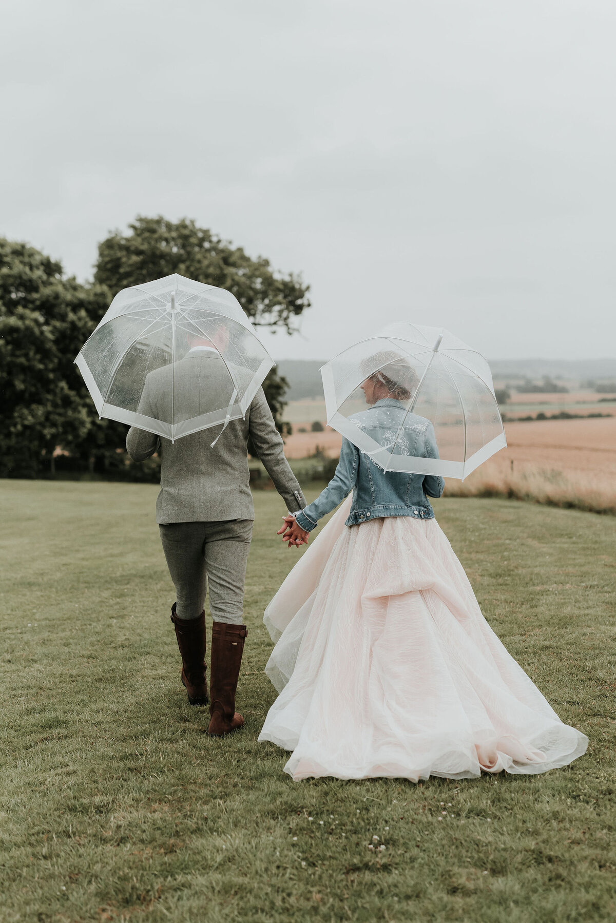 Bride & Groom walking into field with clear umbrellas and wellies at their barn wedding at Odo's Barn, Ashford