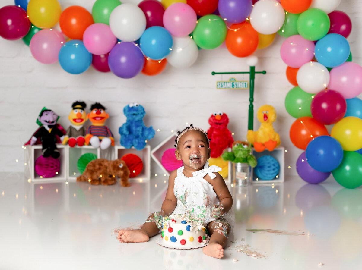 Sesame Street themed cake smash in West Palm Beach and Wellington, FL photography studio. Black baby girl is sitting with a bright polka dot take between her legs, icing is on her hands, legs and face. She has a big smile at the camera. In the background, there is a bright rainbow-colored balloon arch with Sesame street characters and street sign behind her.