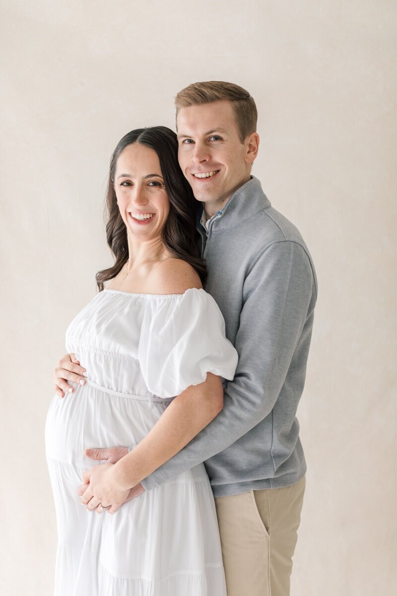 A pregnant woman and her husband stand close together and smile at the camera during their Lawrenceville NJ Maternity Photography session