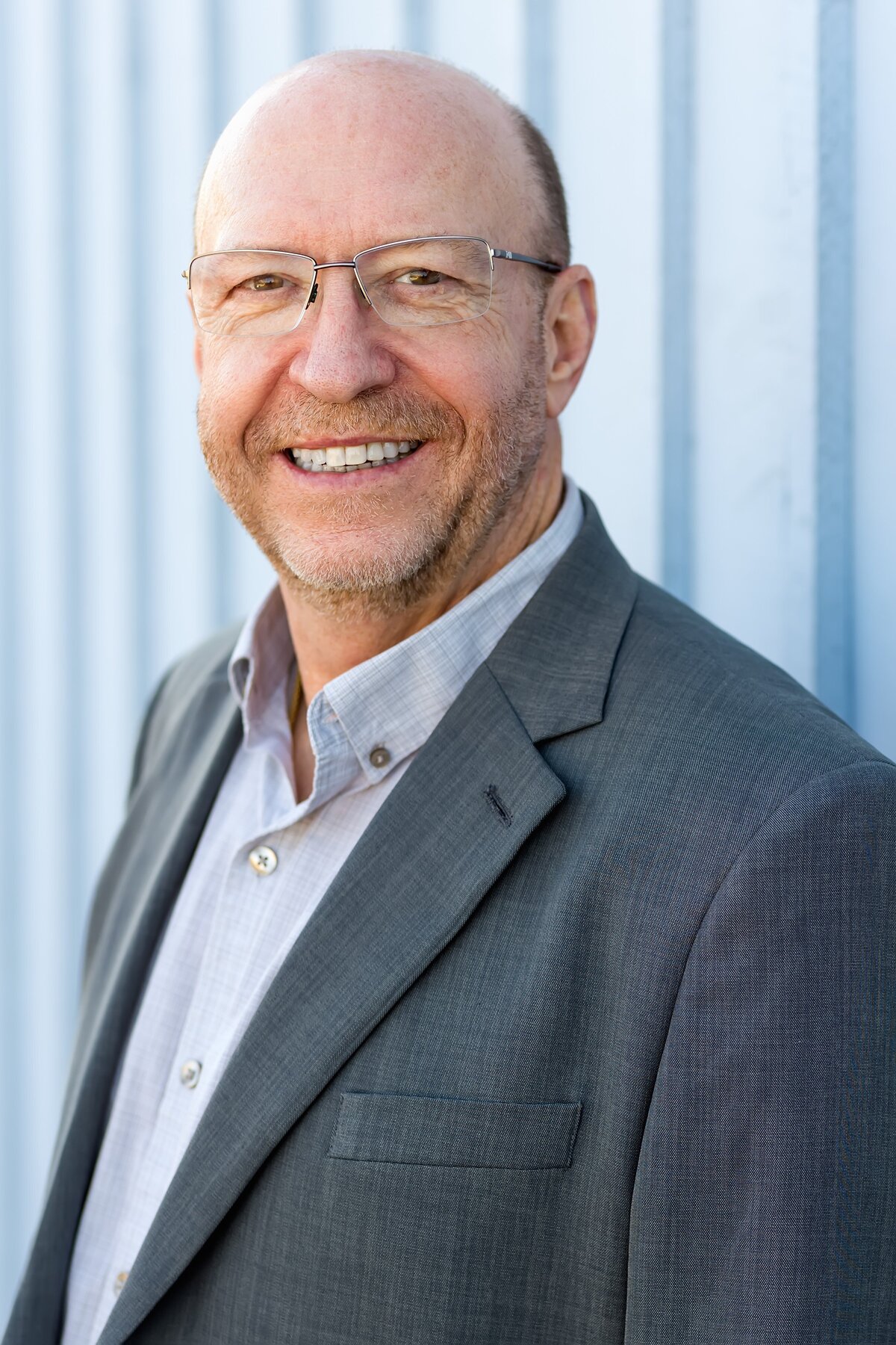 Linkedin headshot of man in suit jacket and wearing glasses while leaning against a blue wall