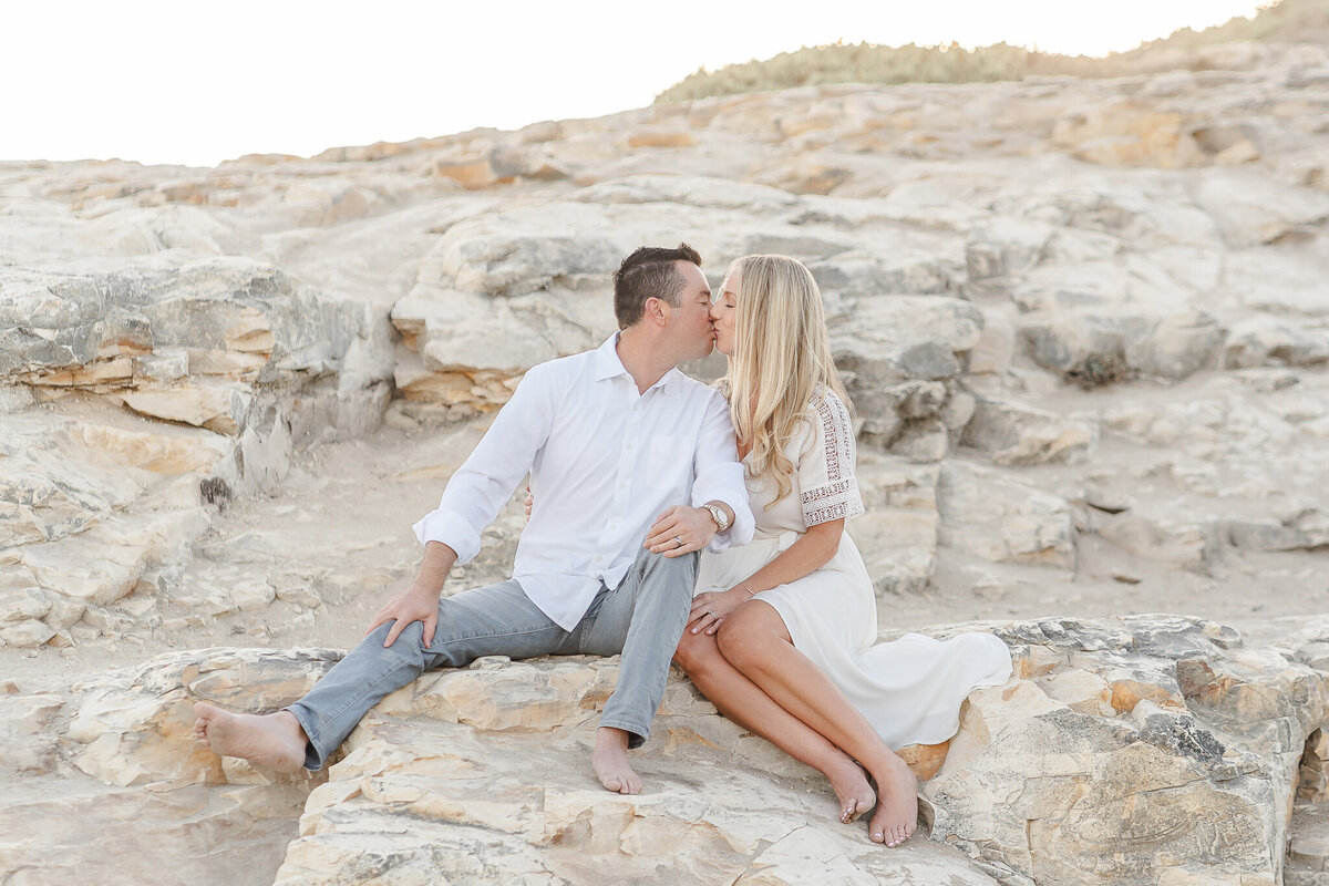 mom and dad wearing white and kissing each other while sitting on a rock at the beach
