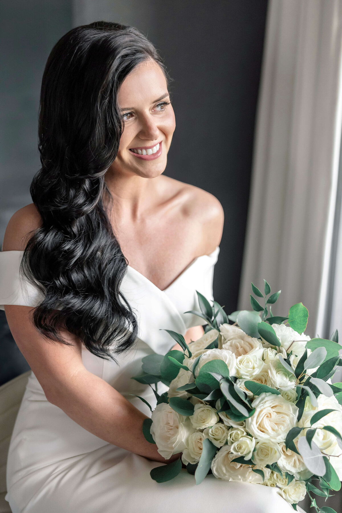 A woman with long dark hair and a white off-the-shoulder dress smiles while holding a bouquet of white roses and green leaves. She is sitting indoors near a window with light filtering in.