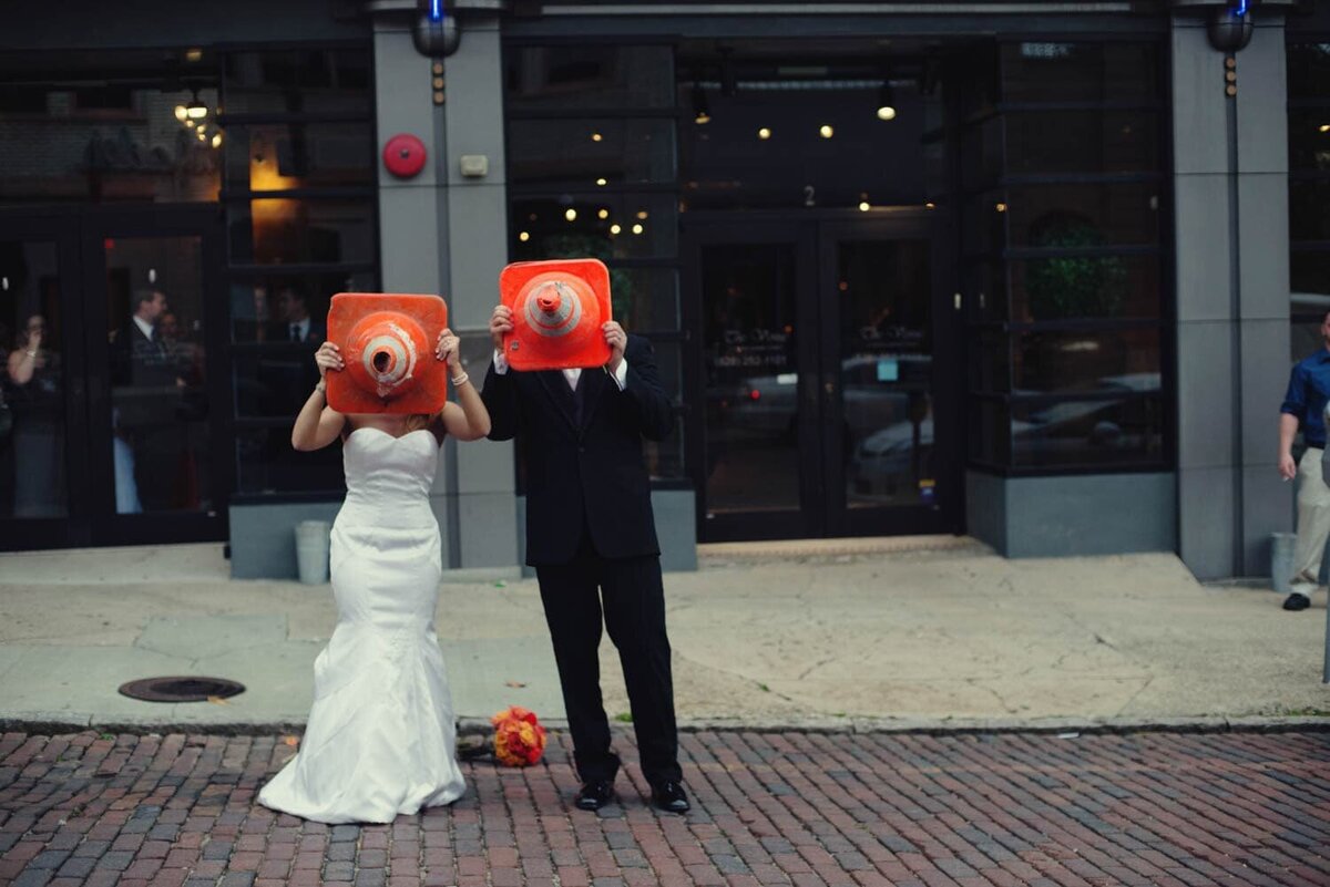 a silly bride and groom hold orange construction cones over their faces