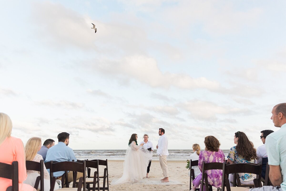 A candid moment during a wedding ceremony on the beach at Crystal Beach, Texas. The bride and groom are holding hands and facing each other in front of their officiant while backed by the ocean. The guests can be seen in the foreground while a seagull flies overhead. The bride is wearing a flowing, white dress while the groom is wearing a white dress shirt and khaki dress pants.