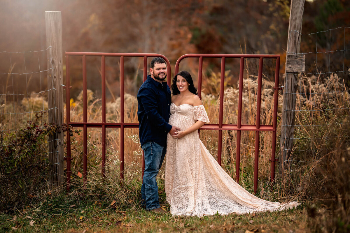A mom and dad  place their hands on the mama's baby bump while standing in front of a red gate in the country