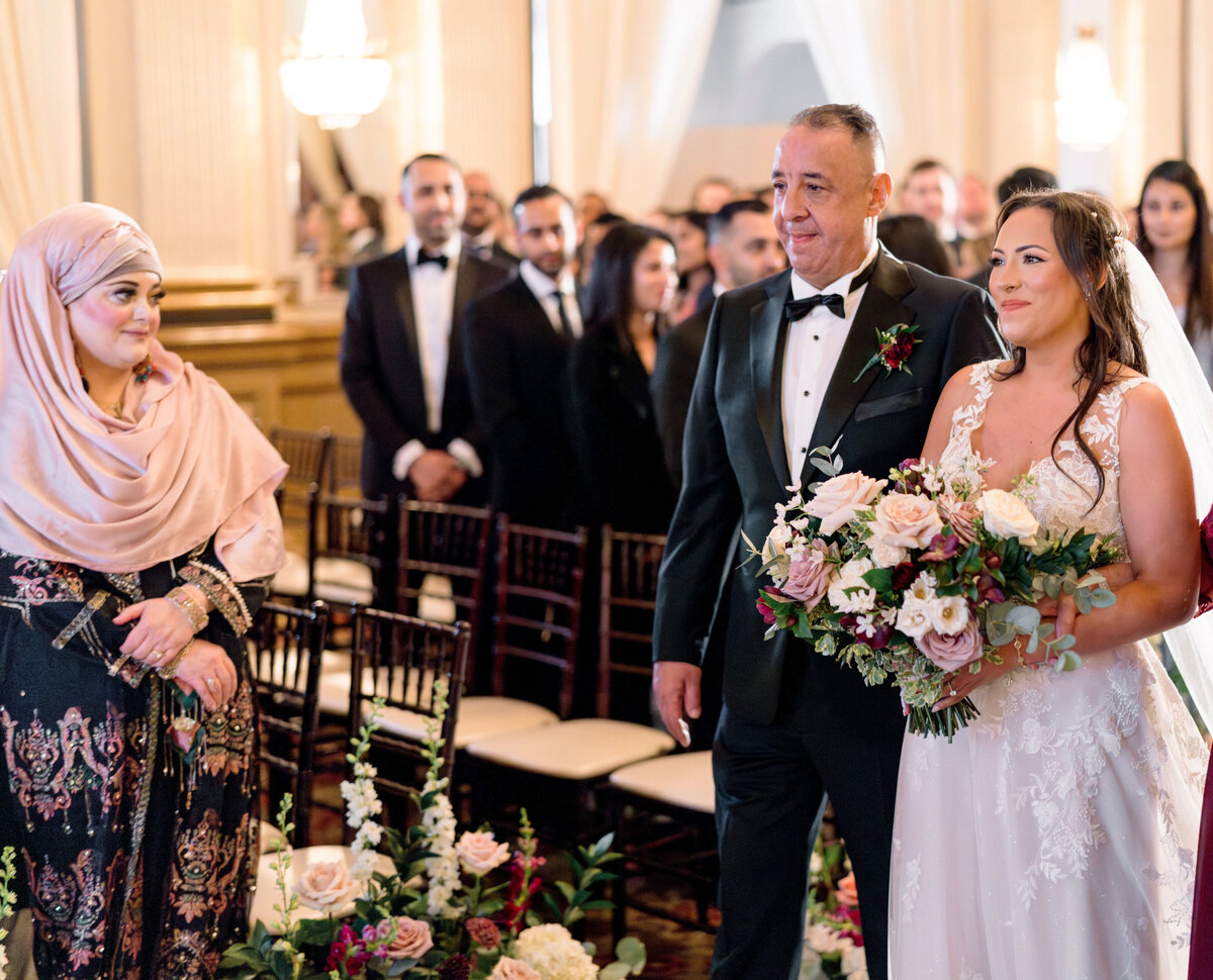 A bride in a white gown holds a bouquet of flowers, walking down the aisle with an older man in a tuxedo. A woman in a headscarf stands nearby, smiling. Guests are seated and watching in the background. The setting is a decorated indoor venue.