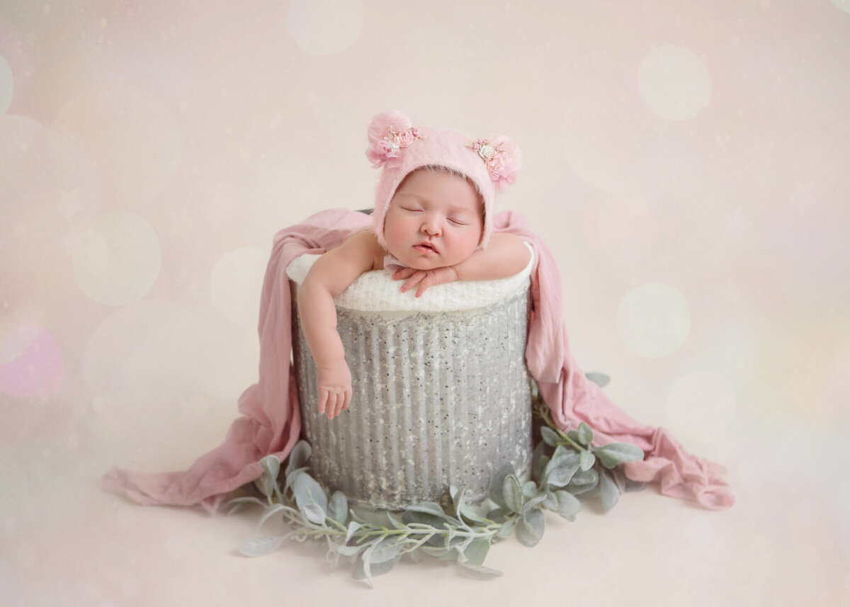 Baby in a pink teddy hat asleep in a bucket with one arm hanging down, taken by Los Angeles baby photographer