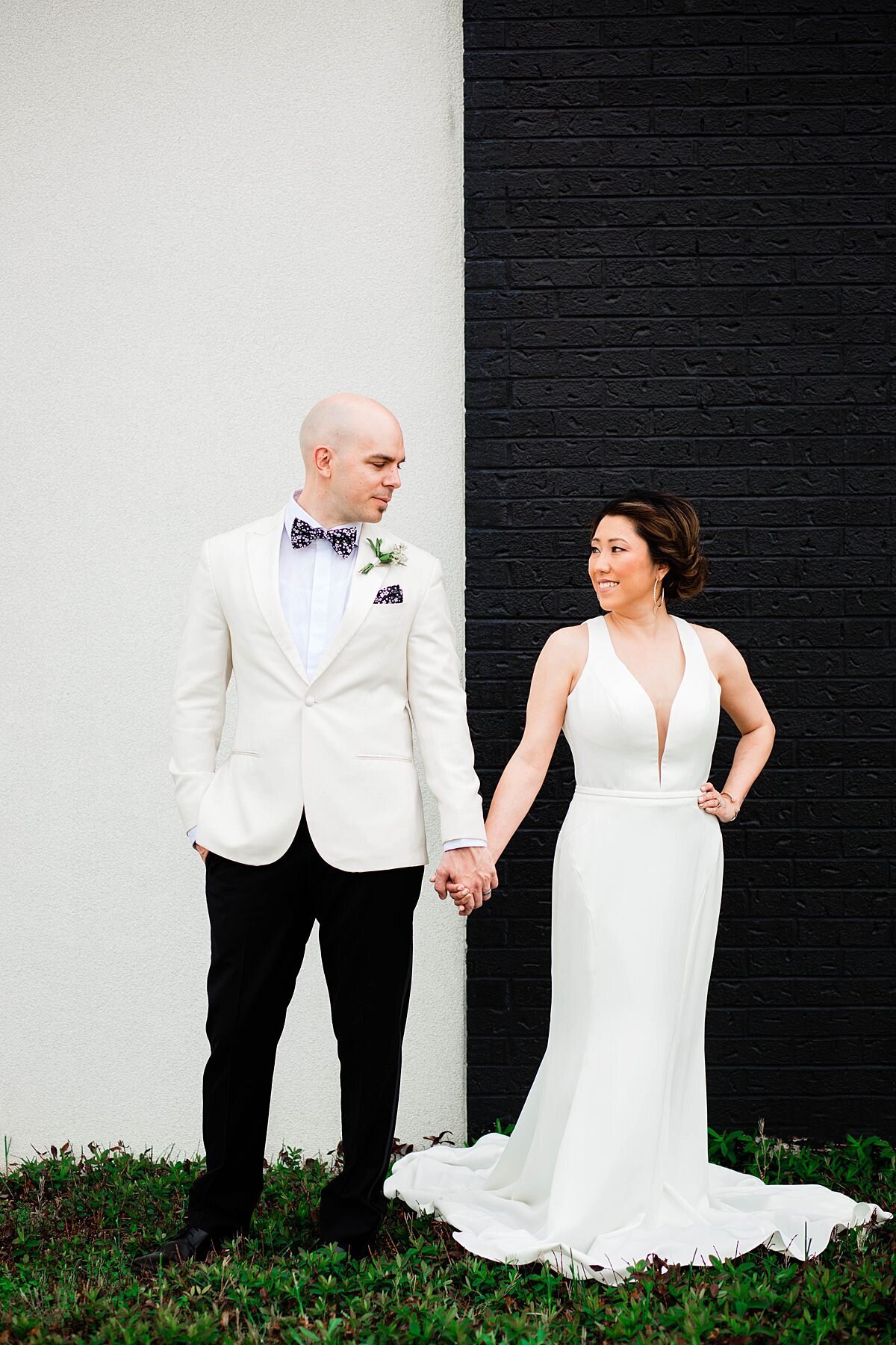 The bride and groom stand outside of the Nashville Ballet. The  Bride stands in front of a black wall wearing a white floor length fitted gown with a long full train and a deeply plunging neckline. She is holding hand with the groom smiling at him. The groom is standing in front of a white wall wearing a white tuxedo jacket and white shirt with black pants a black bowtie with white polka dots and a black pocket square with white polka dots. He has a white boutonniere pinned to his lapel . He is holding hands wit the bride smiling down at her.