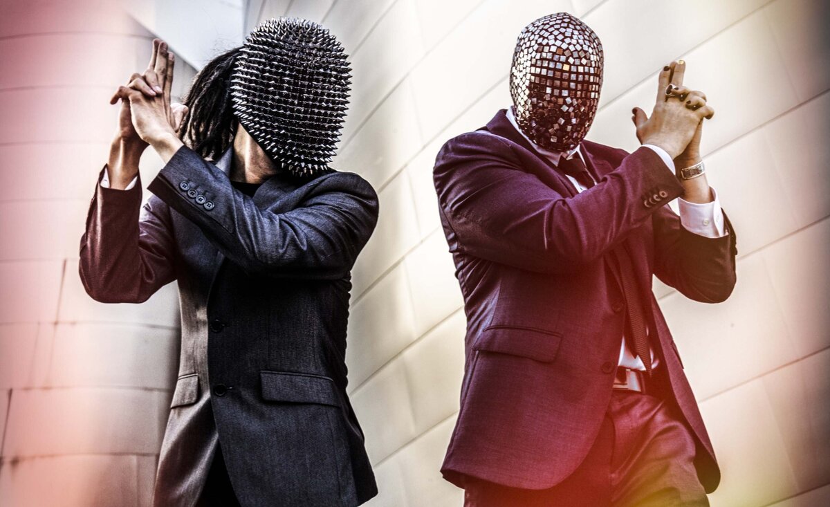 Musical duo portrait Casper and Linboy wearing suits with masks holding hands up like guns
