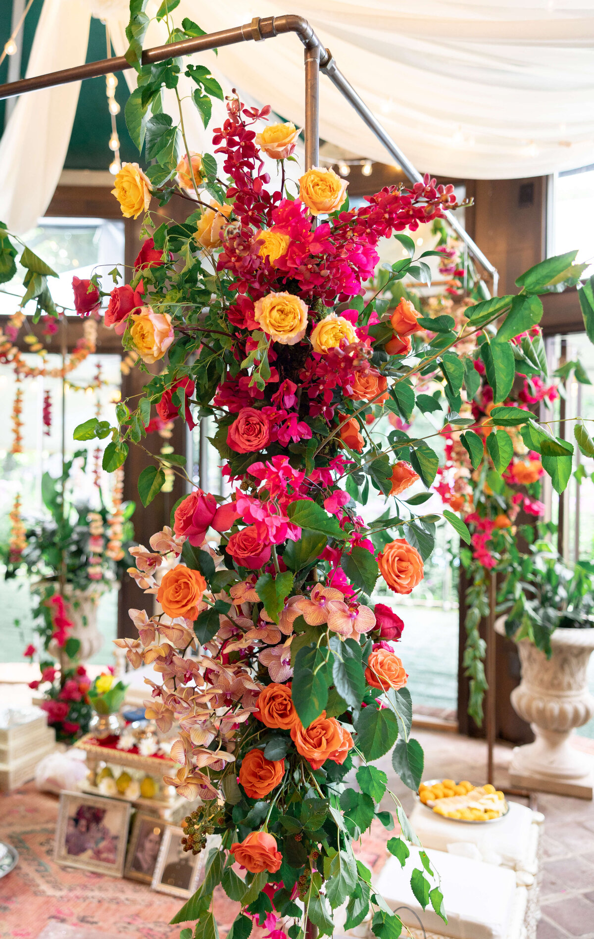 A vibrant floral arrangement drapes over a metal frame, featuring an array of red, yellow, pink, and orange flowers with lush green leaves. In the background, more flowers and a partially visible table with decorative items can be seen.