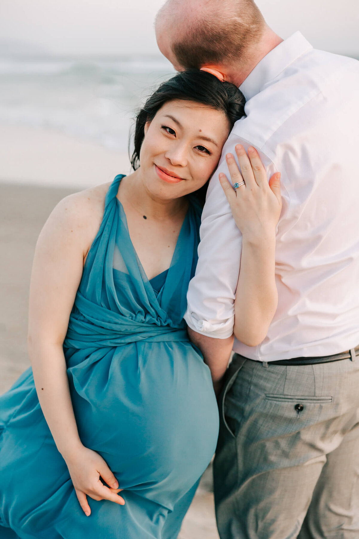 mom holding husband in beach maternity photograph for wearing blue while husband wears white shirt and grey pants