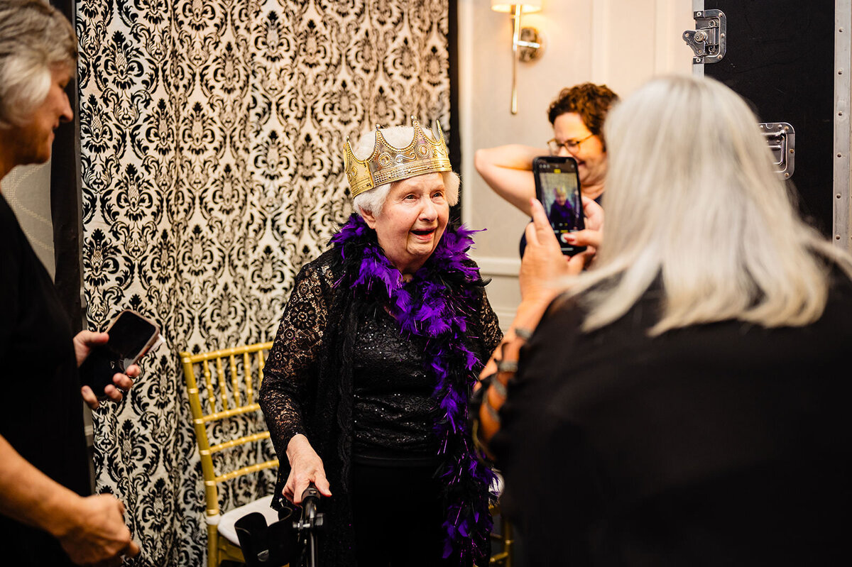 An elderly woman wearing a crown and a purple feather boa is photographed, with a person taking her photo in the background