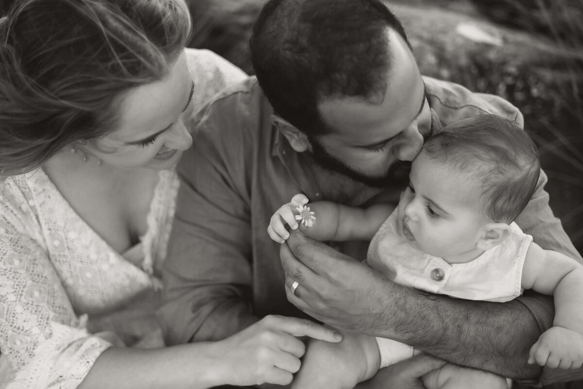 Baby s holding onto a flower as dad kisses his head and mum cuddles dad and touches baby's thigh.