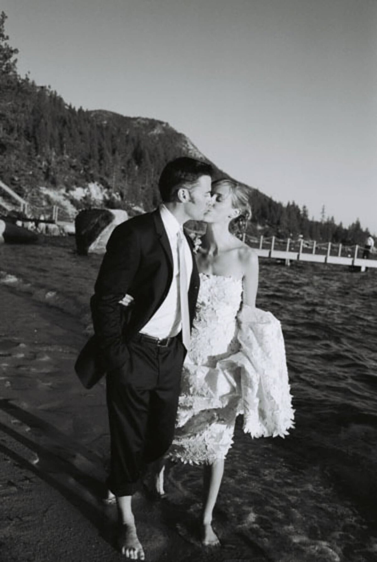 Newlyweds walking on the beach in Lake Tahoe feet in water bride holding her dress as they are kissing. Black and white film image