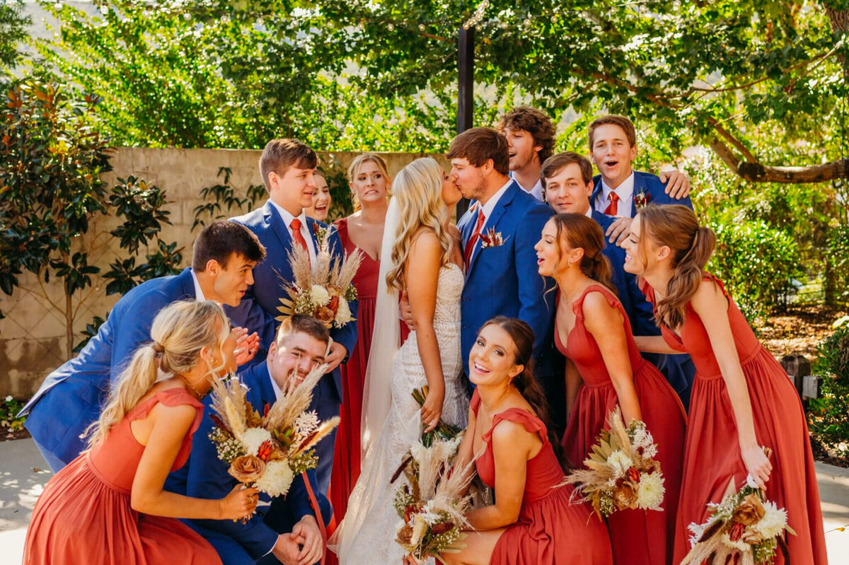 Photo of a bride and groom kissing while their wedding party surround them in a group hug