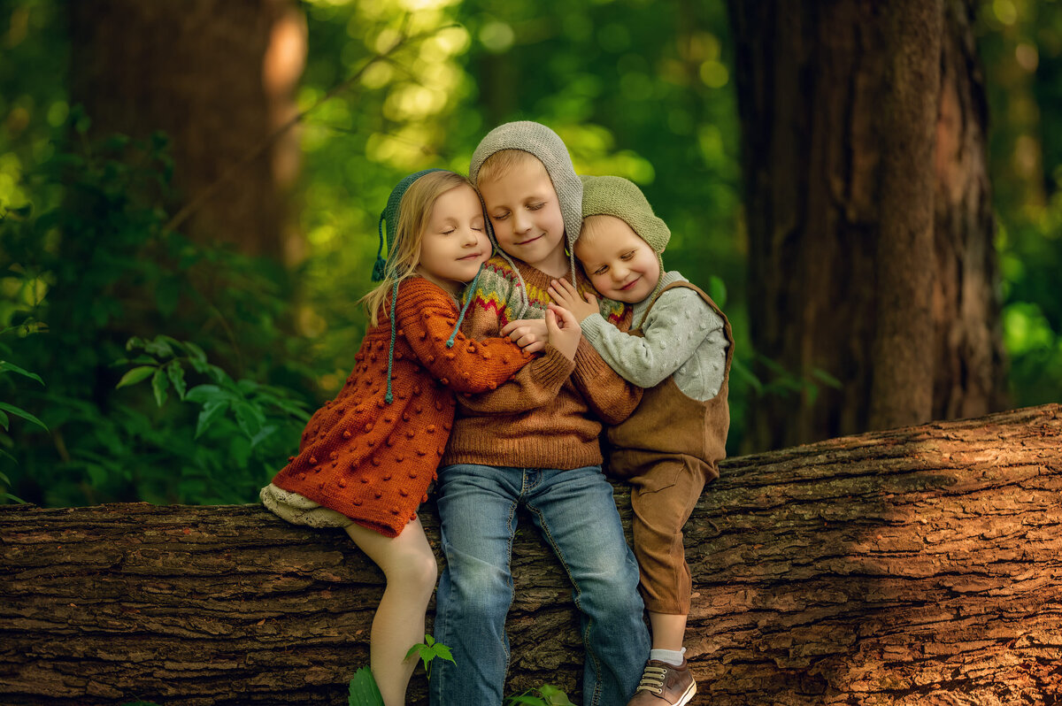 Three siblings embrace on a large log in a bright, ethereal forest portrait session by Kara Reese of Waukesha, WI.