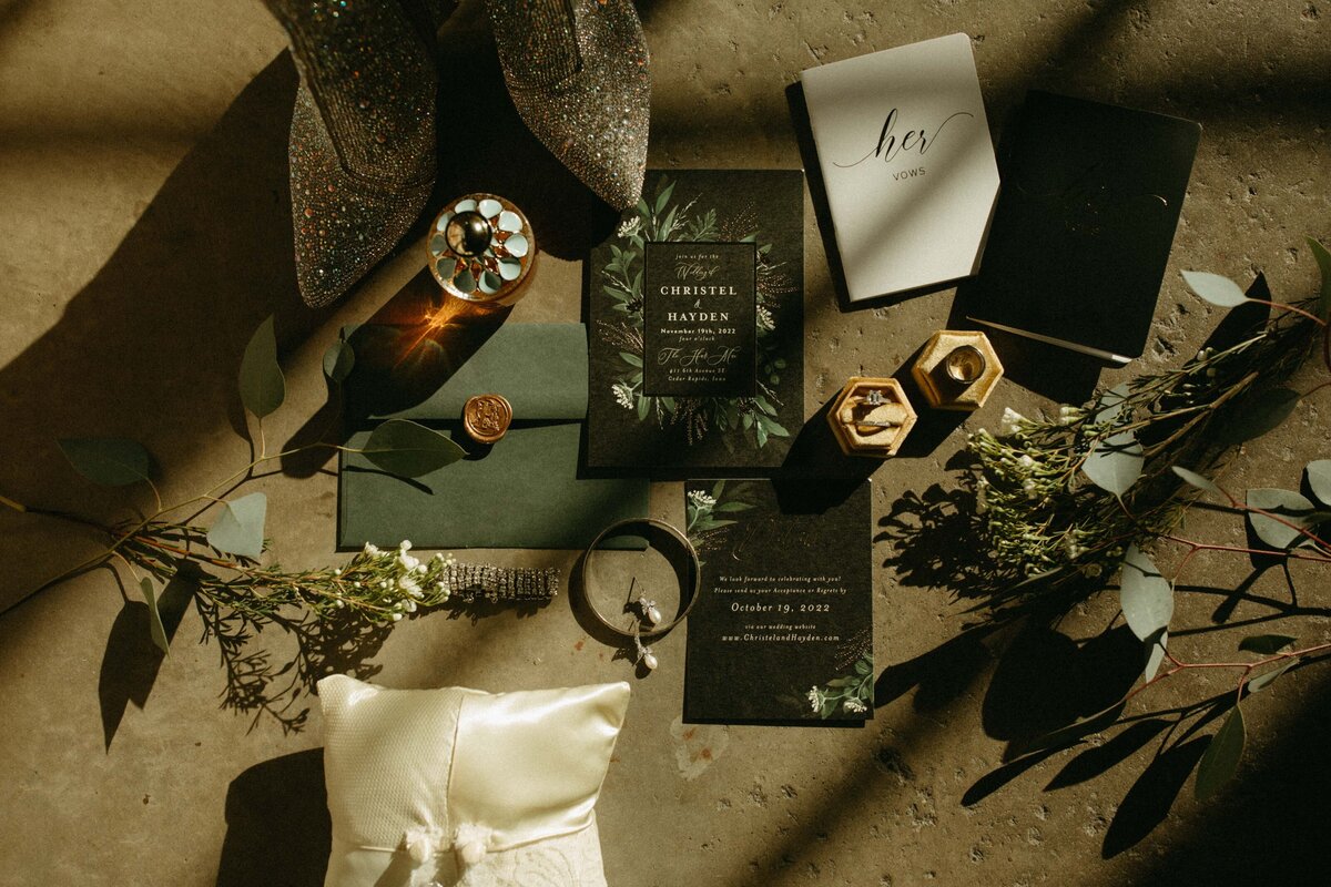 Flat lay of wedding accessories including invitations, shoes, rings, and decorative greenery on a textured surface with sunlight casting shadows for an Iowa wedding.