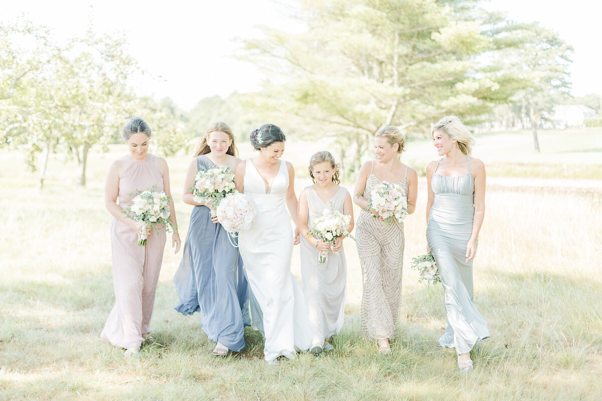 Bride is walking with bridesmaids and junior bridesmaids in a Boston park for casual wedding photo. They are all smiling and looking at each other. Captured by Boston wedding photographer Lia Rose Weddings