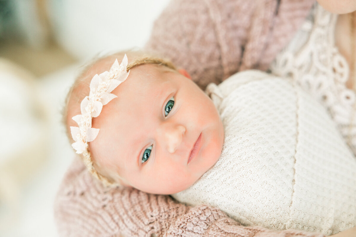 baby girl looks right at camera while being held by mom, wearing a pink bow and cream wrap.
