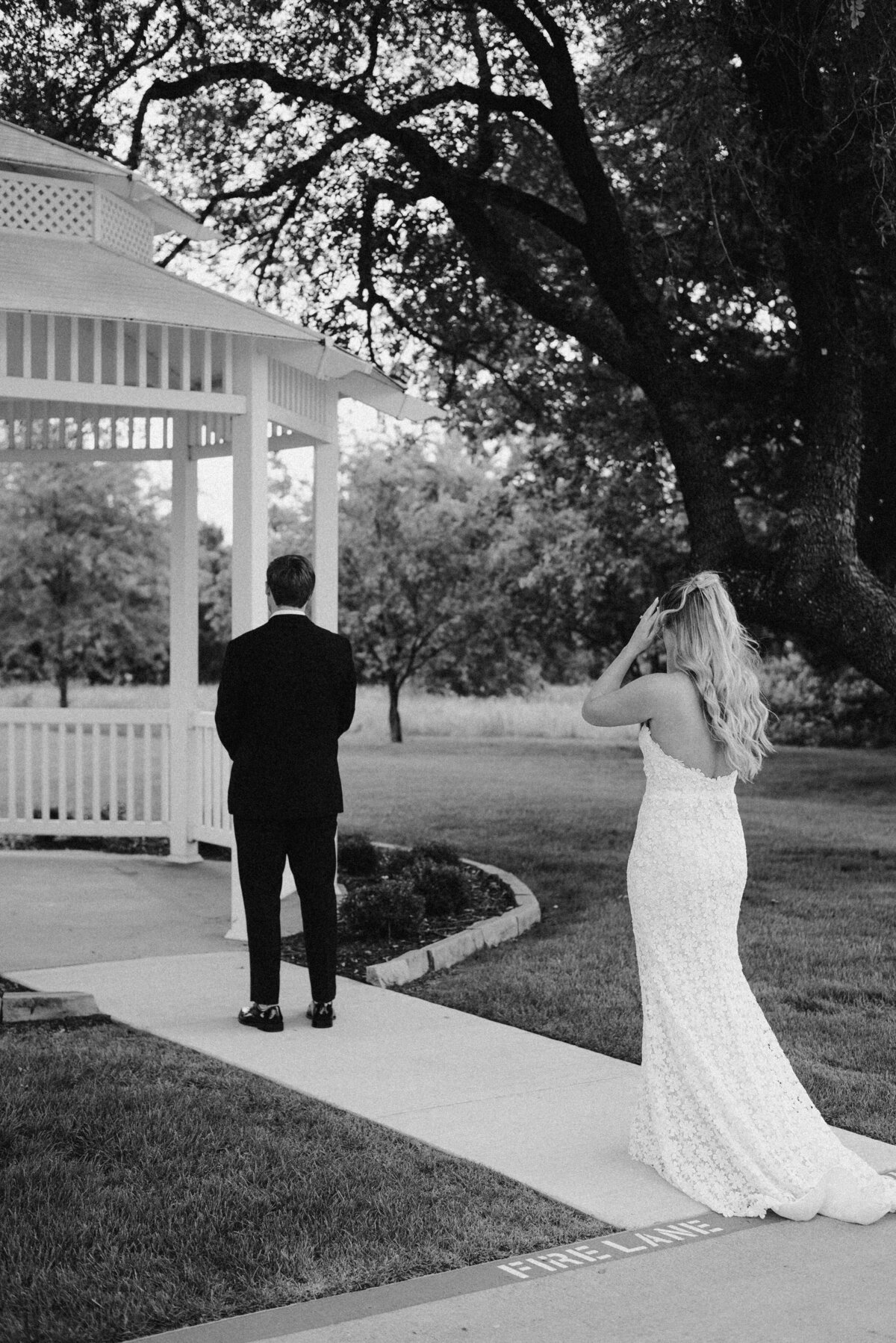 A bride in a lace gown approaches a groom standing near a gazebo on a lush garden path. The scene is serene and intimate, captured in black and white.