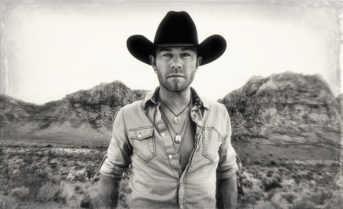 Country musician portrait black and white Aaron Pritchett wearing denim shirt black cowboy hat mountain range background