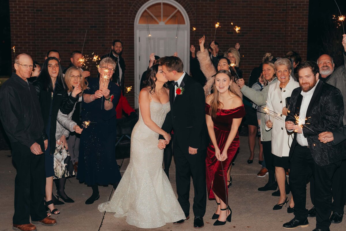 Newlywed couple kissing outside a church at night, surrounded by family and friends holding lit sparklers after their Iowa wedding.