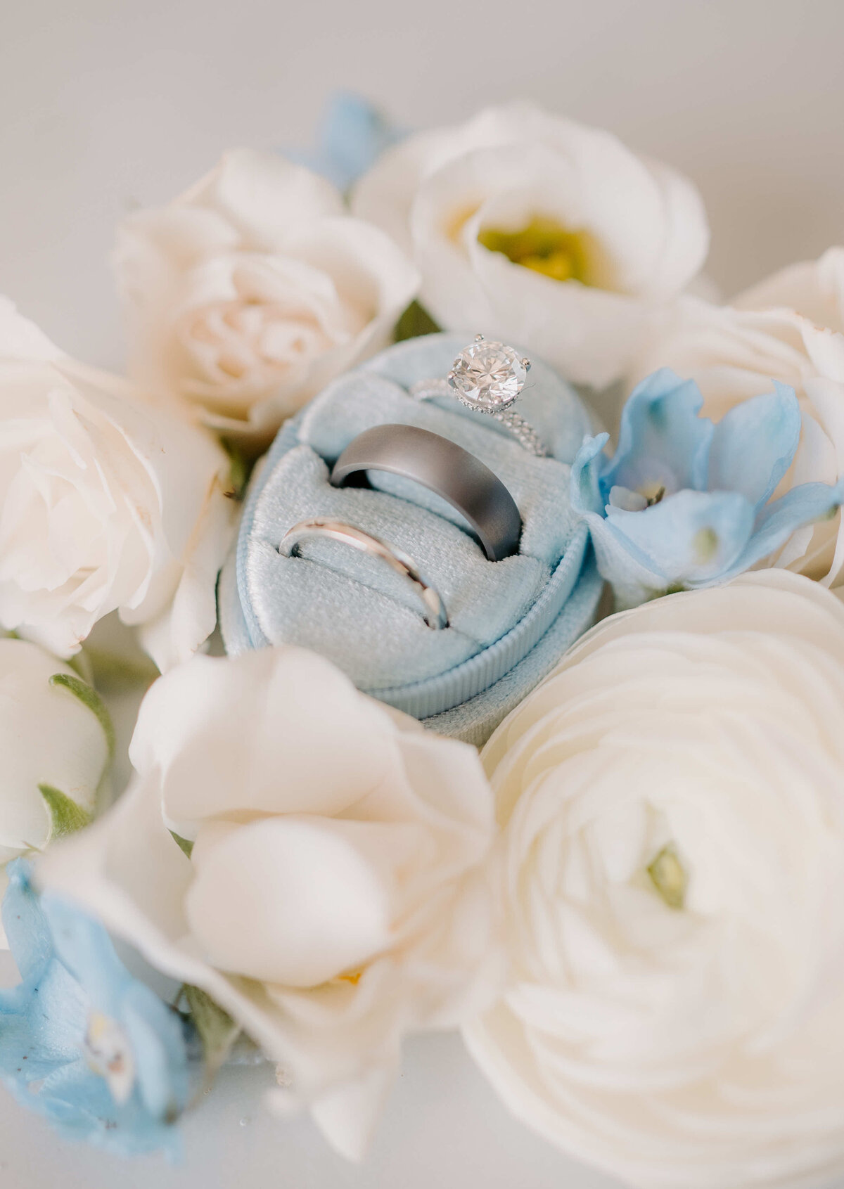 A velvet ring box with a diamond ring and two bands is nestled among soft white and blue flowers, creating a romantic and elegant scene.