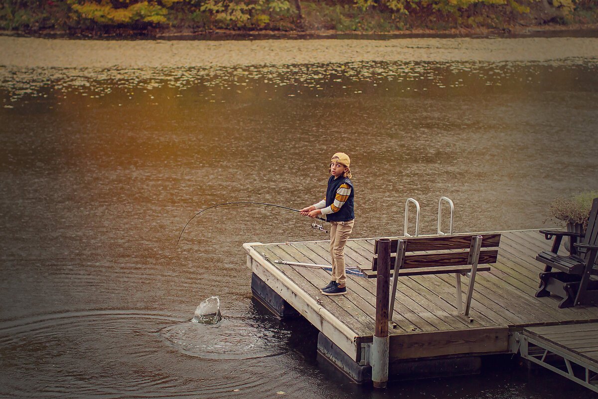 Boy is fishing off the dock with a fish on the line. He's wearing a yellow cap, blue vest, and there are fall colors all around.