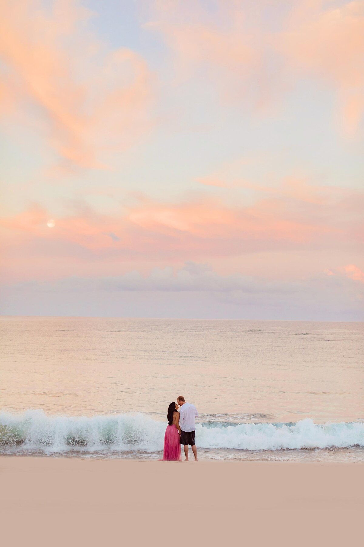 Wide shot of a couple kissing at sunrise as a full moon sets behind them on the beach in Wailea
