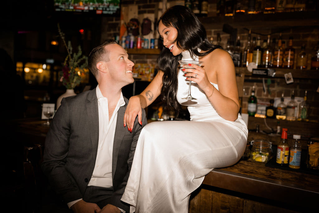 bride sitting on a bar holding a drink while she leans on her groom's shoulder