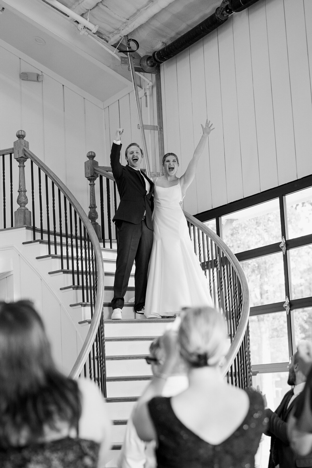 A couple making their entrance to their reception at a wedding at Bella Cavalli Events in Aubrey, Texas. The groom on the left is wearing a dark suit with white shoes, is holding the bride around the waist, is holding his other hand up in celebration, and is cheering. The bride on the right is doing the same except for wearing a white wedding dress. Guests can be seen in the foreground looking up at the couple as they descend a staircase.