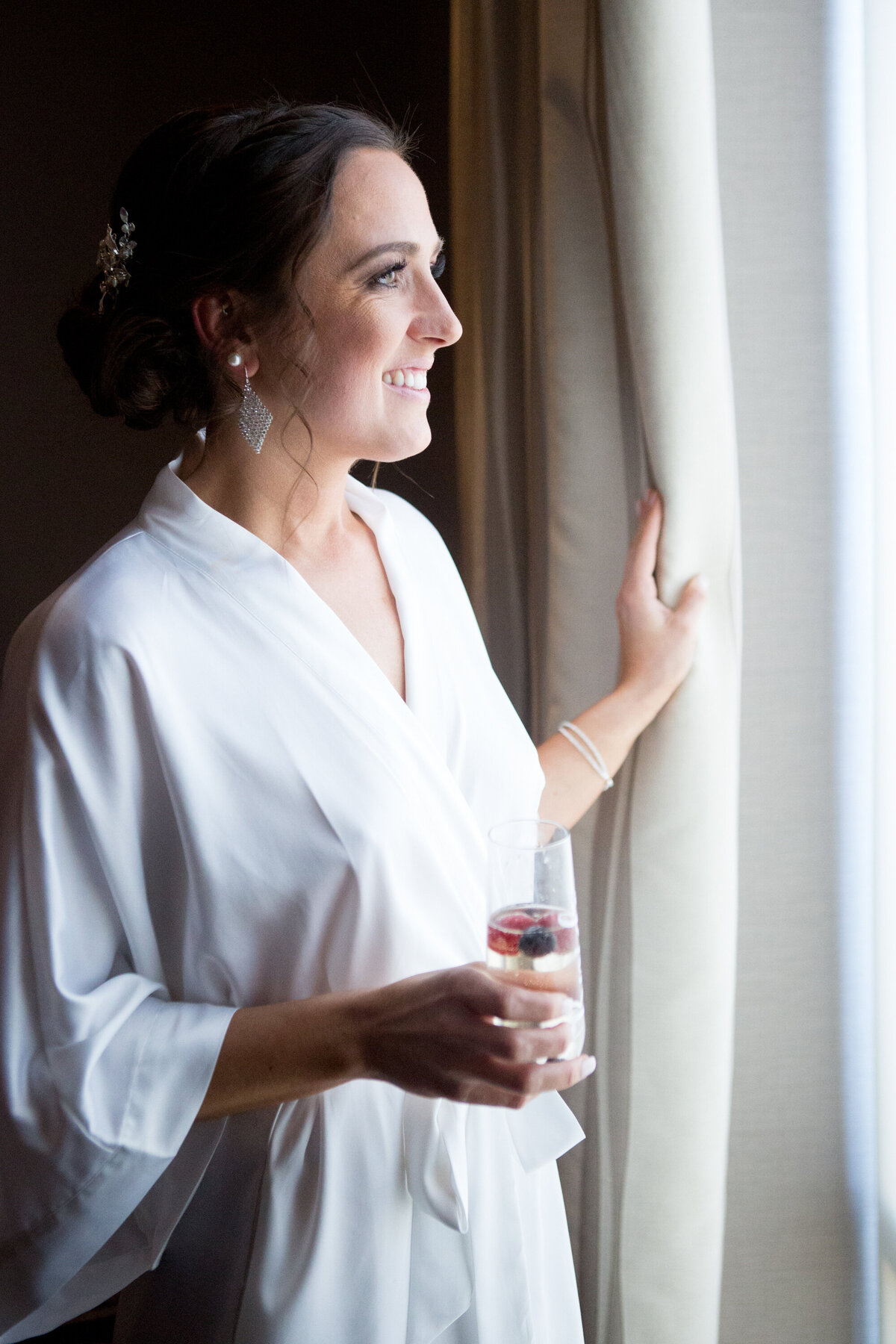 A woman in a white robe looks out the window holding a champagne glass Pearl River Hilton wedding photographer Milestone Images