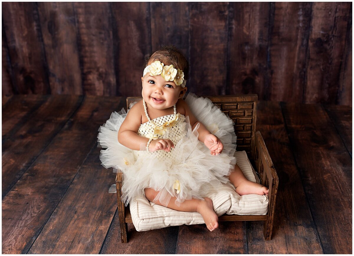 Six month old baby girl sitting on a little prop bench and looking on the camera smiling.