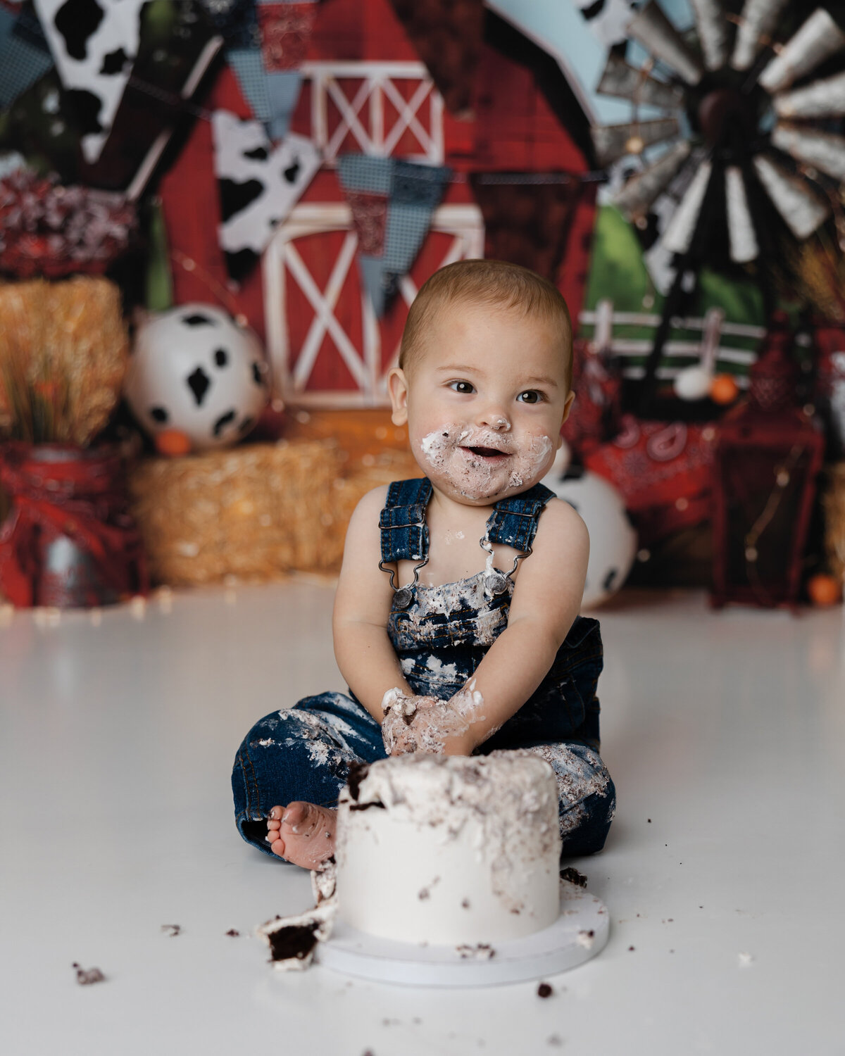 Smiling baby boy in denim overalls smashing a white frosted cake during his farm-themed first birthday session.
Photograph by Yucaipa cake smash photographer Kaitlyn Dawn Photography.