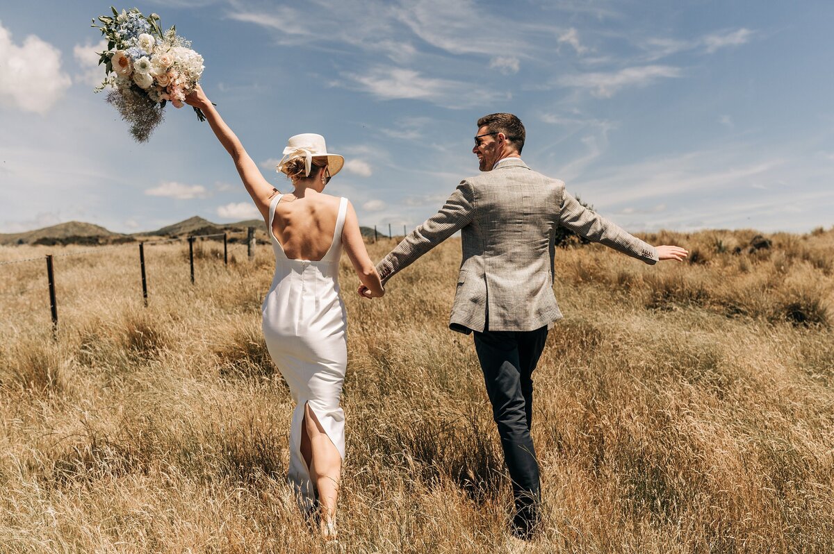 bride and groom walking away in long dry grass wither hills blenheim with white hat holding pastel bouquet and tweed blazer in summer sun helicopter