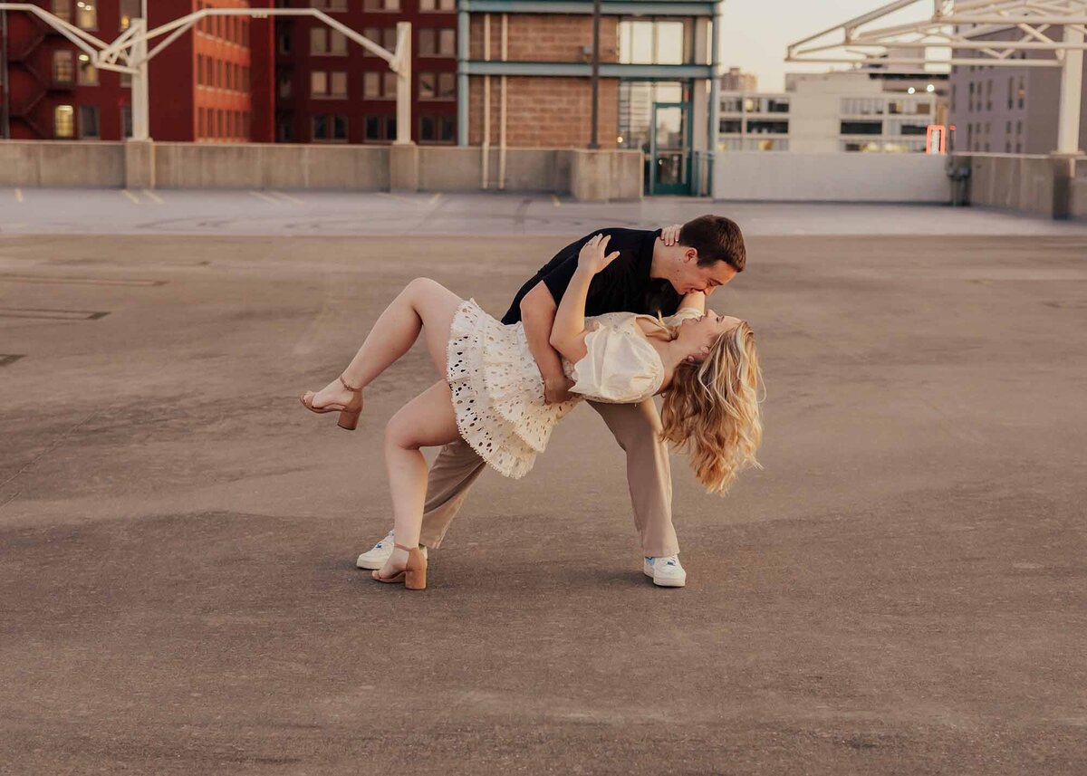 Maddie Rae Photography wide angle shot of a couple dip kissing on the top of a parking garage. she has one leg off the ground