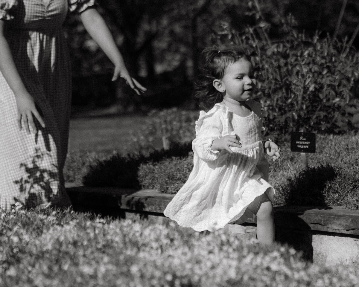 Toddler running and laughing with mother’s arms following close, photographed naturally in a garden setting in Columbia, Missouri, capturing real connection and joy.