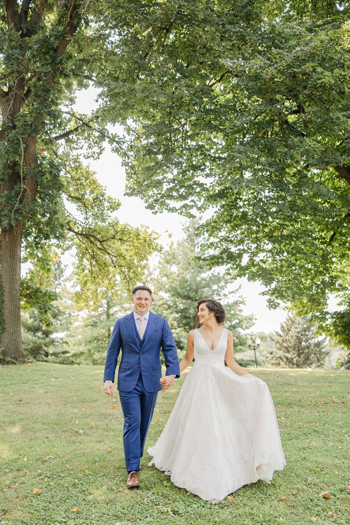A bride in a white dress and a groom in a blue suit walk hand in hand through a grassy park in Iowa, smiling joyfully.