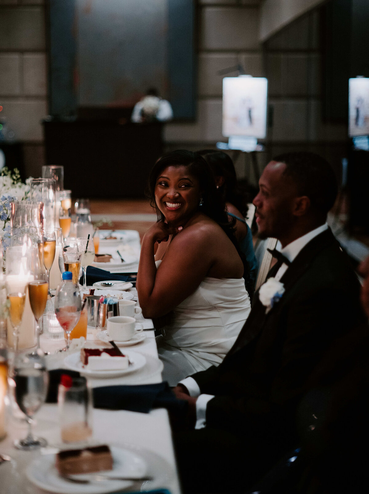 A woman in a white dress sits at a table, smiling warmly at a man in a tuxedo during a formal event. The table is elegantly set with drinks and plates.