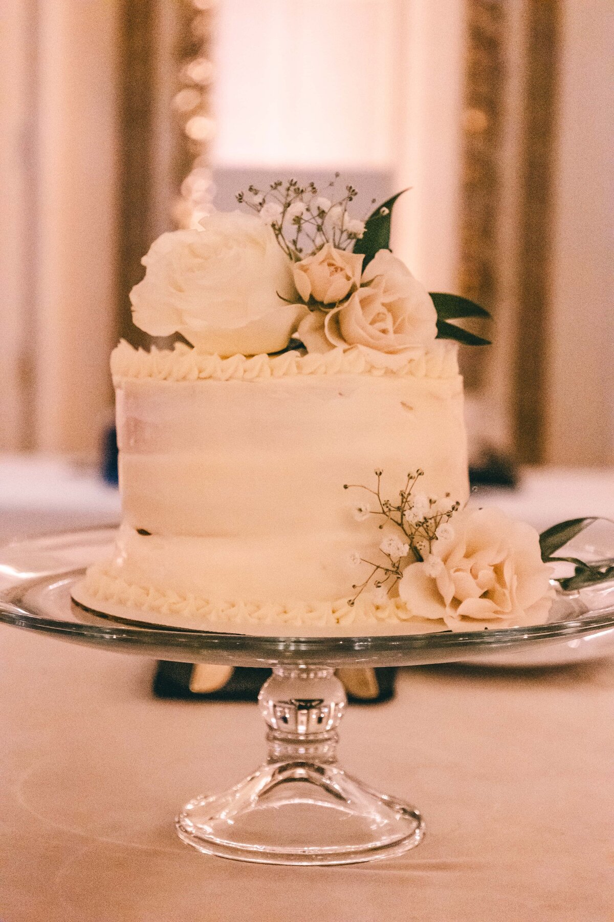 A two-tiered wedding cake adorned with white roses and small flowers on a glass cake stand, set in a dimly lit room at a park farm winery wedding.