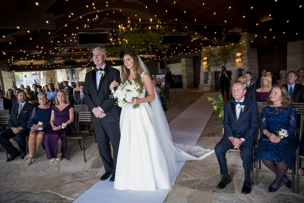 A bride and her father stand at the end of the aisle waiting to be given away with twinkle lights on the ceiling.
