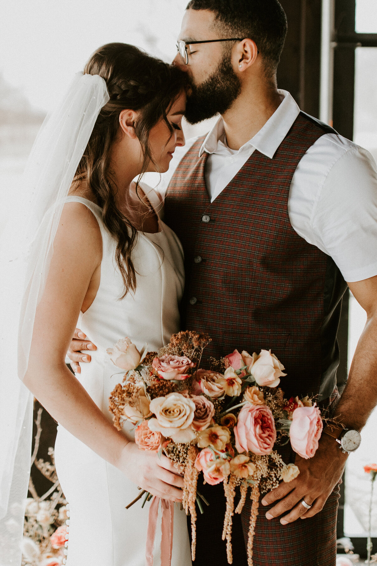 A groom wearing a brown suit kisses bride wearing a white wedding gown on the forehead while she holds a bouquet.