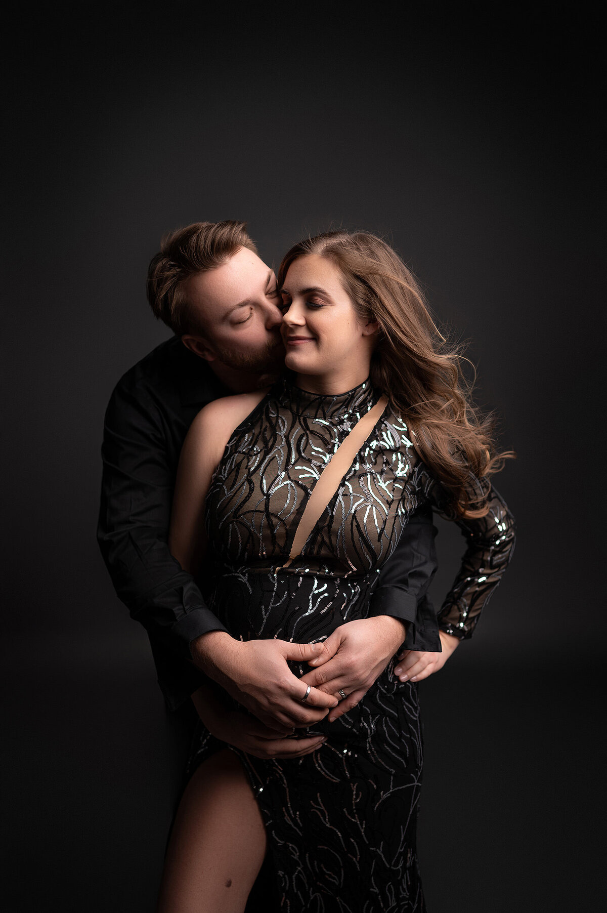 Pregnant woman in edgy black maternity gown looks to the side while her husband hugs her from behind and kisses her cheek.