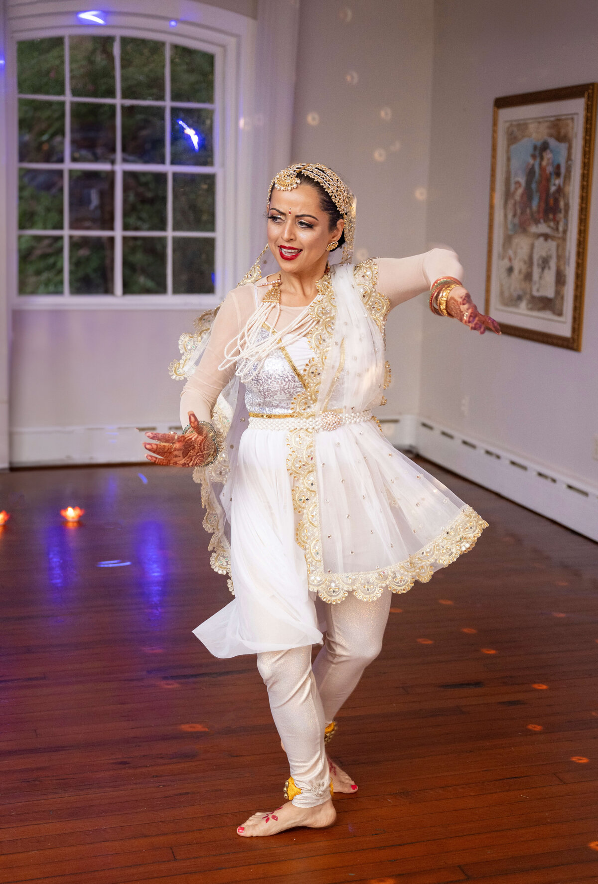 A person in traditional dance attire performs classical Indian dance in a room with wooden floors, a large window, and wall art. They wear a white and gold outfit, with intricate jewelry and decorated hands.
