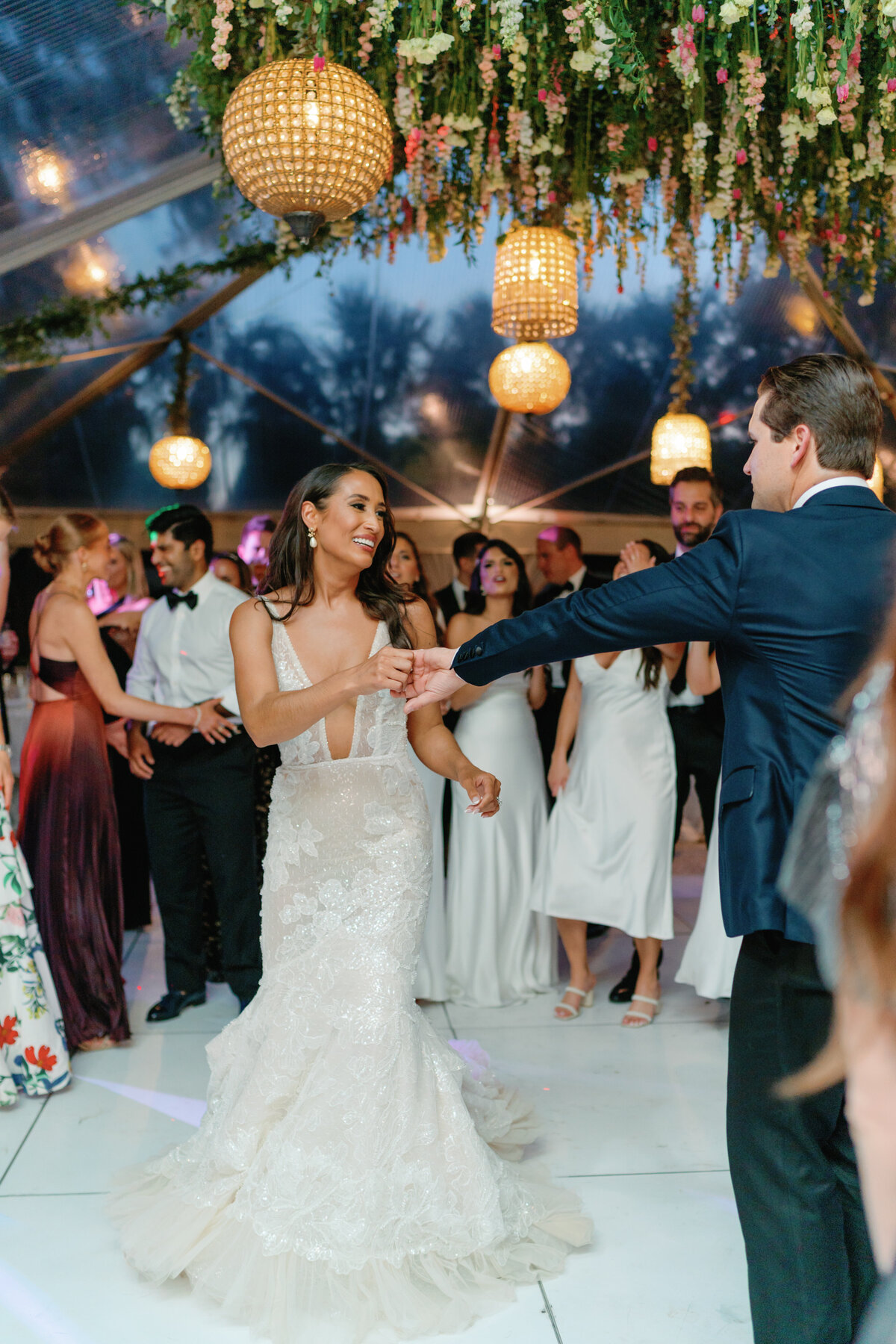 Dark haired bride with plunging neckline Berta wedding dress on all white dance floor with groom in the crowd. Hanging birdcage lighting and upside-down florals.  Spring Thomas Bennett House wedding in Charleston.