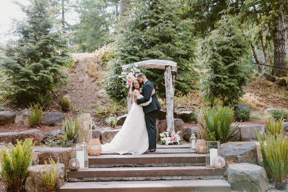 An image from Portland Wedding Photographers of newlyweds snuggling under a log arbor in a garden